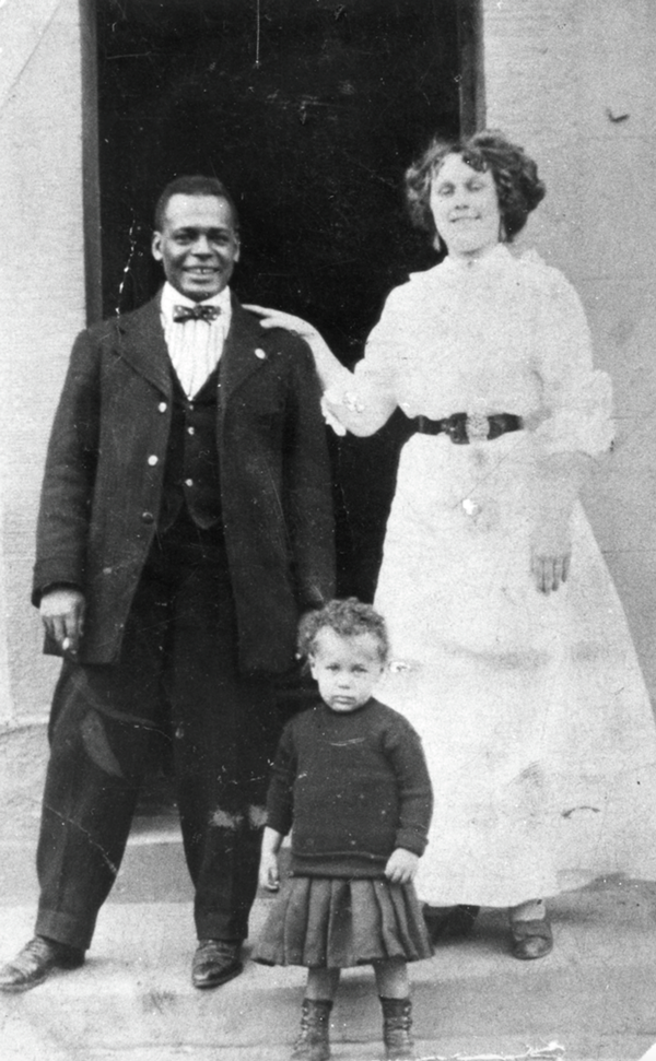 photo of a black man and a white woman in Edwardian dress, standing on the front steps of a house with a small child in front of them. The woman is slightly taller and has her hand resting affectionately on the man's shoulder. They're both smiling.